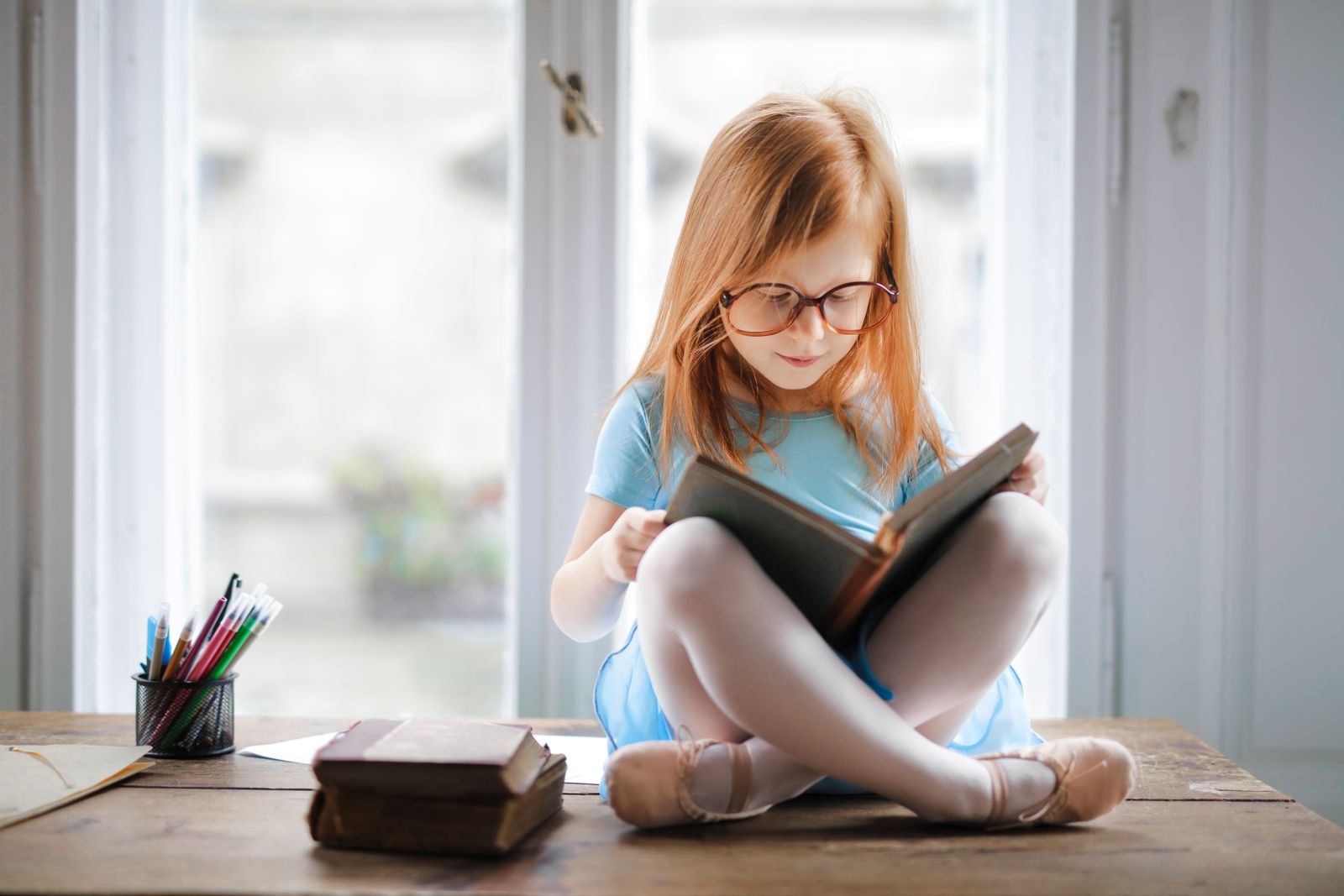 Дівчинка в окулярах читає книжку біля вікна